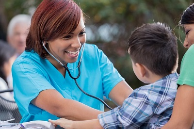 Beautiful mid adult African American nurse or doctor smiles while checking a little boy's heartbeat. The nurse is volunteering at a free outdoor clinic. She is wearing a stethoscope and scrubs. The boy's mother is holding him.