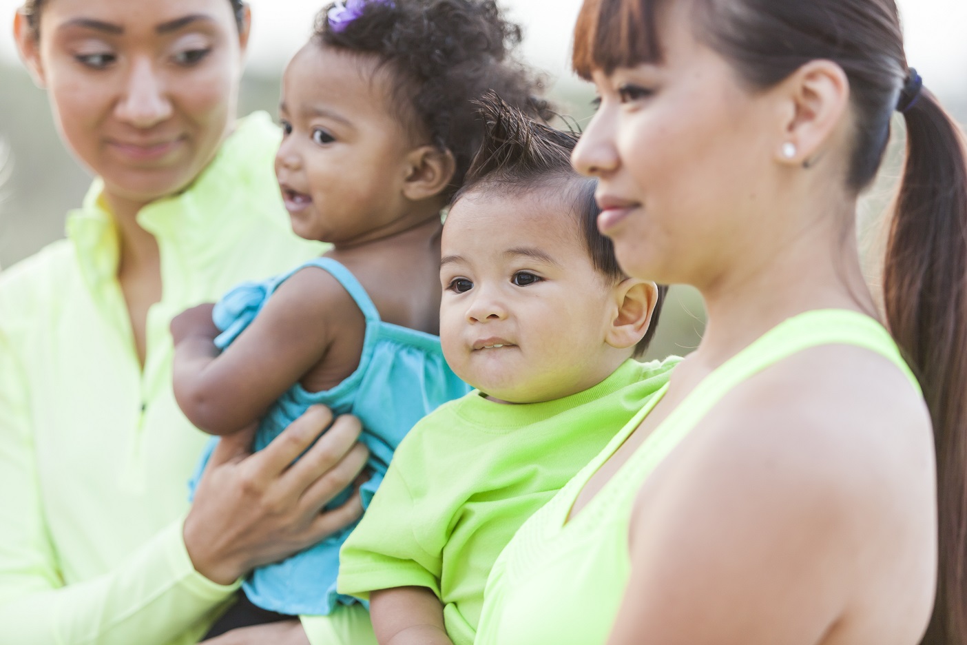 Close up of two multi-ethnic mothers carrying their babies. The focus is on the mixed race (Asian and Caucasian) baby boy in the light green shirt. He is relaxed in his mother's arms.